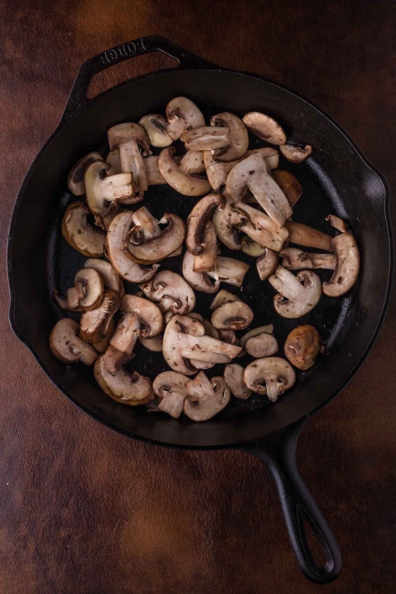 Sliced mushrooms cooking in a black cast iron skillet on a dark brown surface.