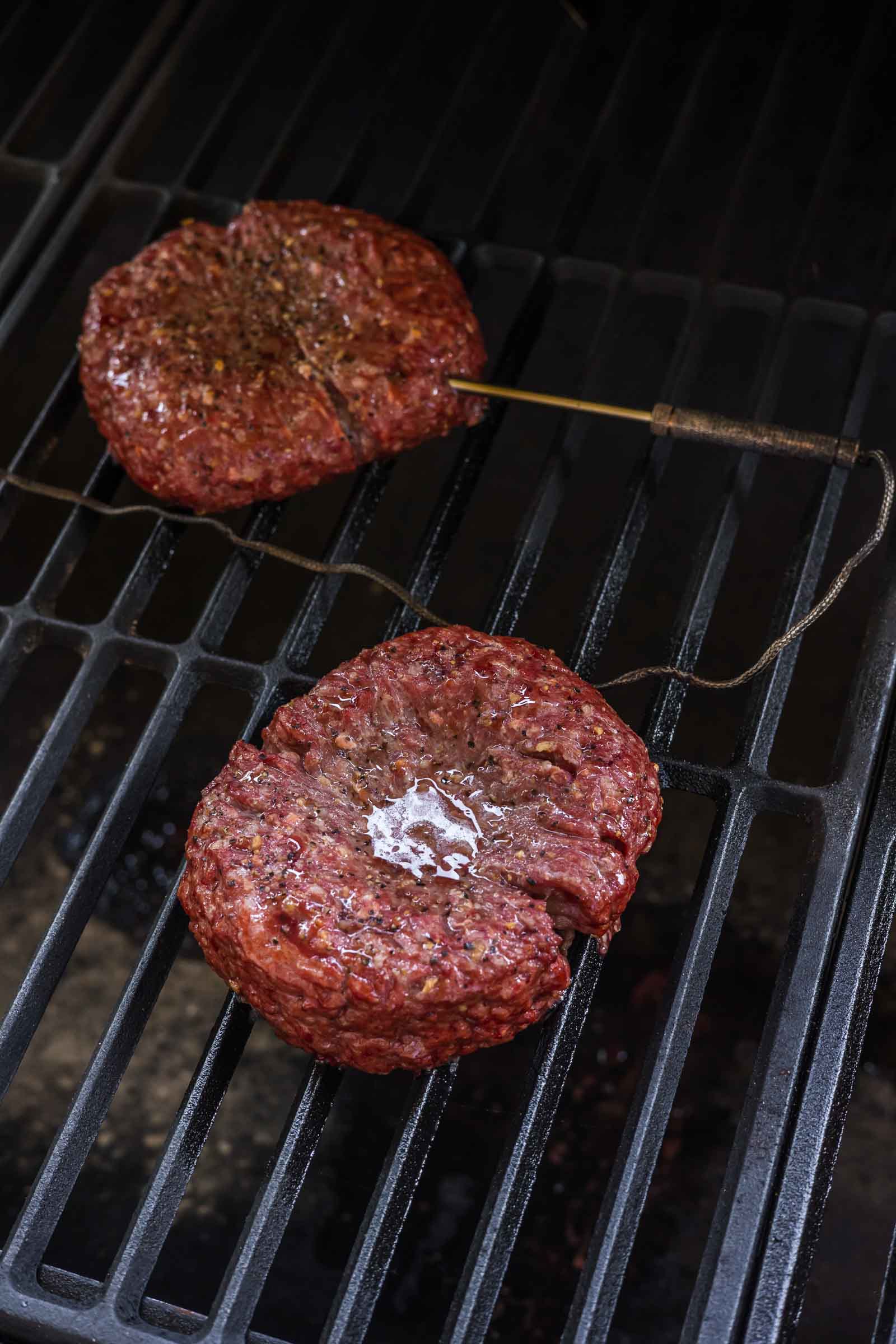 Two raw burger patties on a grill, one with a meat thermometer inserted, and a small pat of butter melting on top of the other.