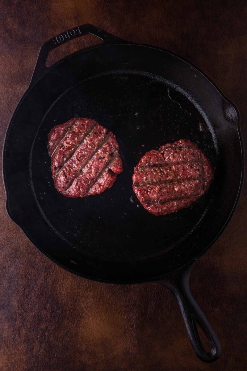 Two raw hamburger patties with grill marks are placed side by side in a black cast iron skillet on a dark brown surface.