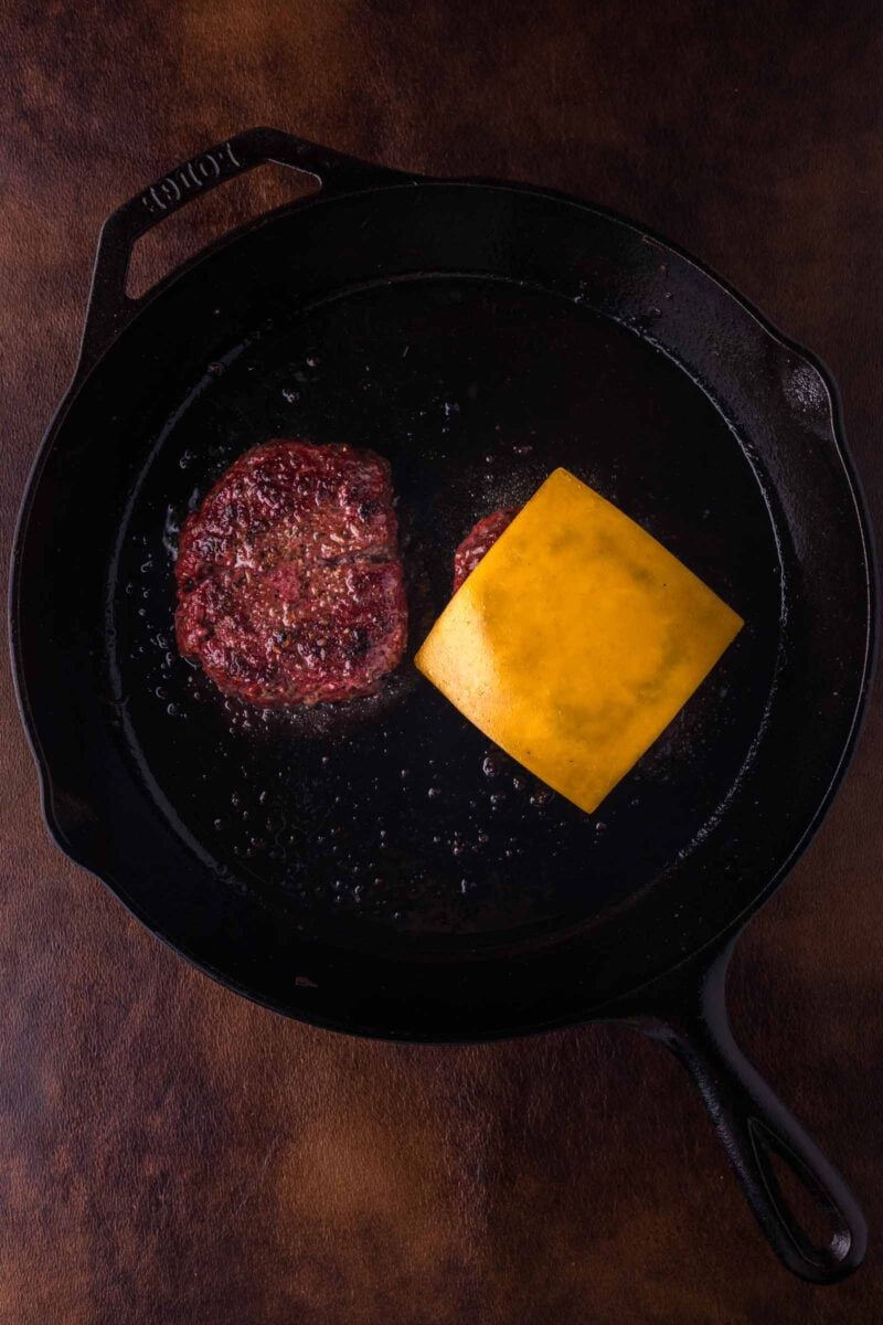 A hamburger patty with a slice of cheddar cheese melting on top cooks in a black cast iron skillet.