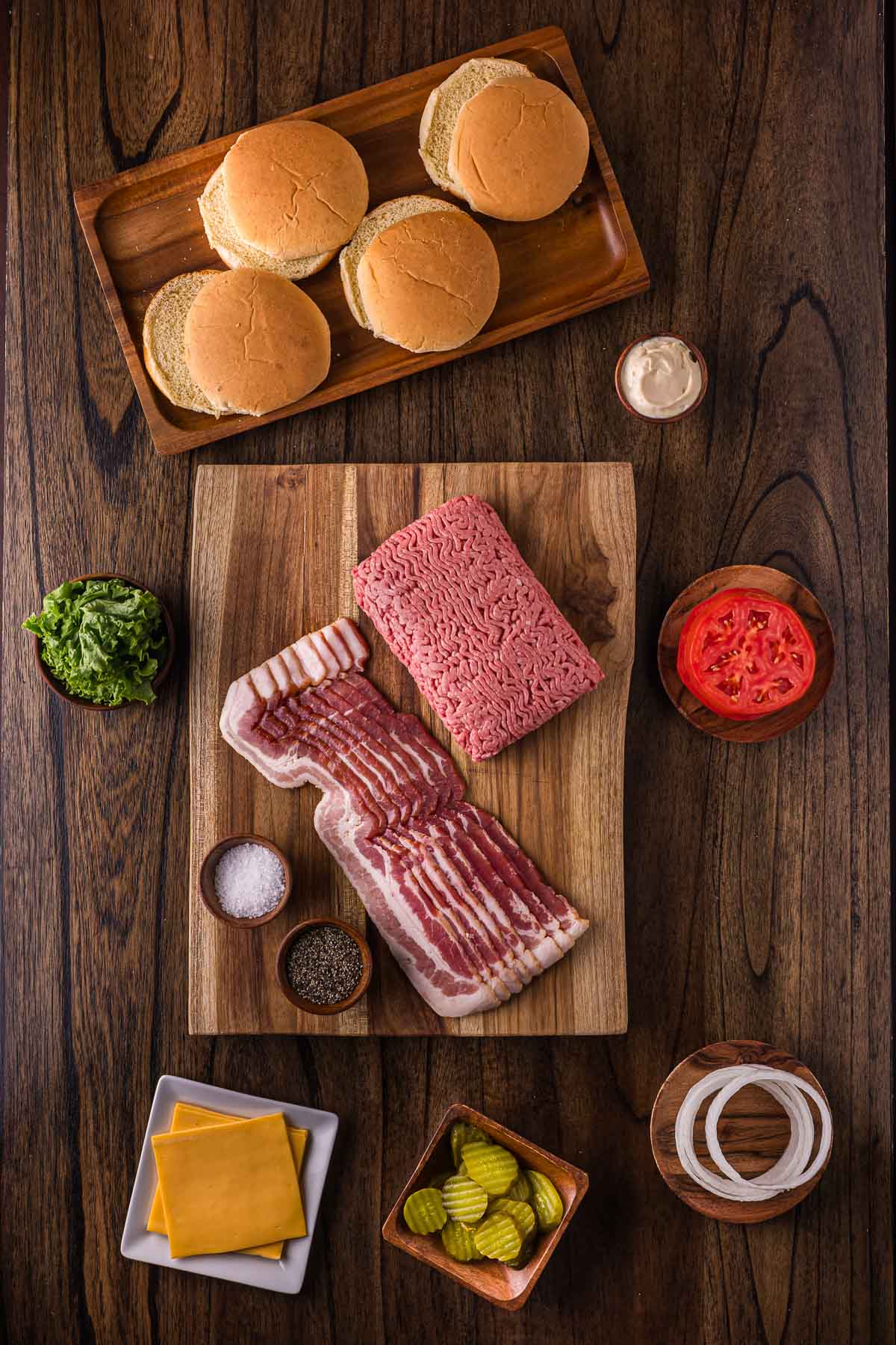 Top-down view of burger ingredients on a wooden table: buns, ground beef, bacon, lettuce, tomato, onion, pickles, cheese slices, salt, pepper, and a small cup of sauce.