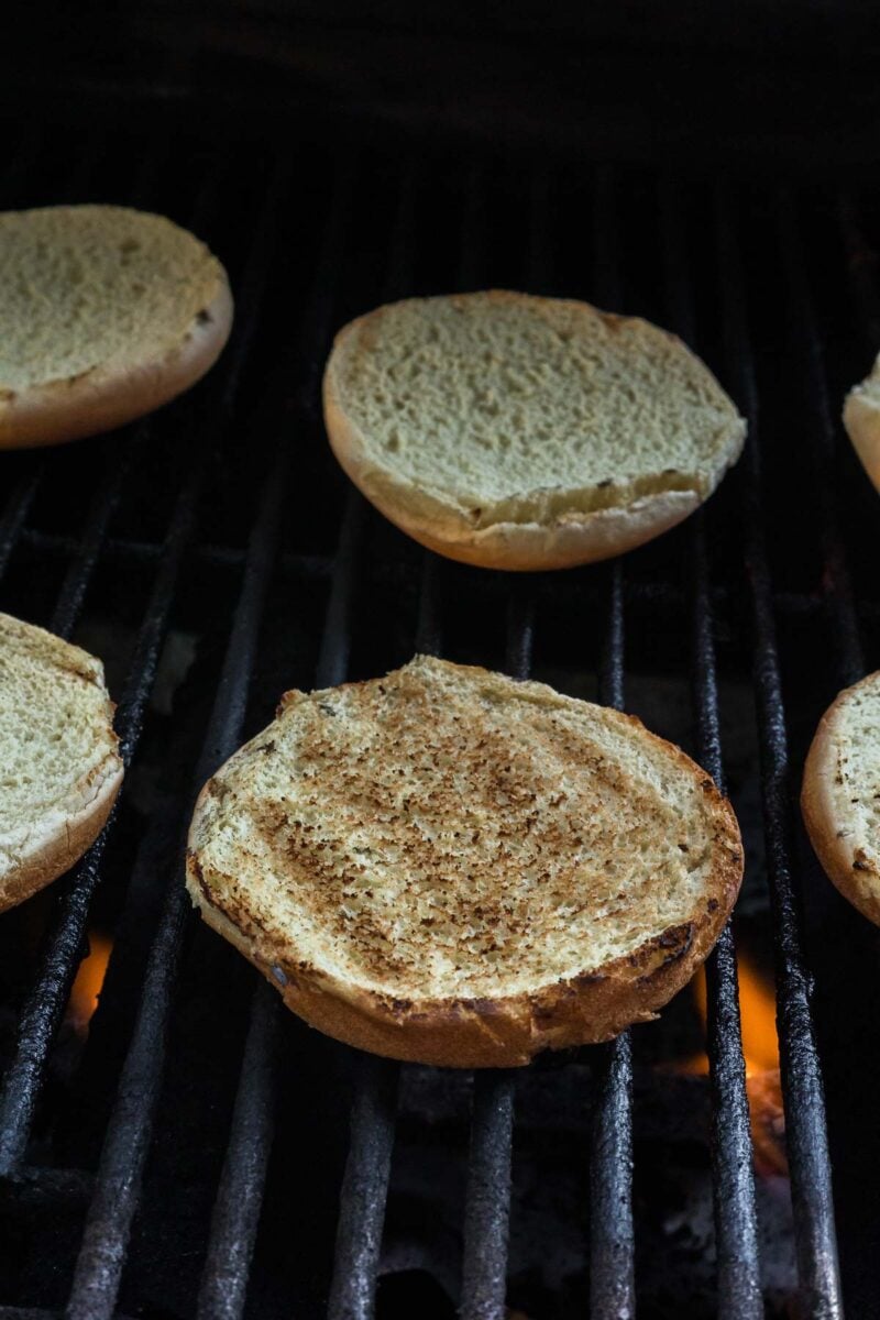 Several hamburger bun halves are toasting on a grill, with visible grill marks and a slight char from the heat below.