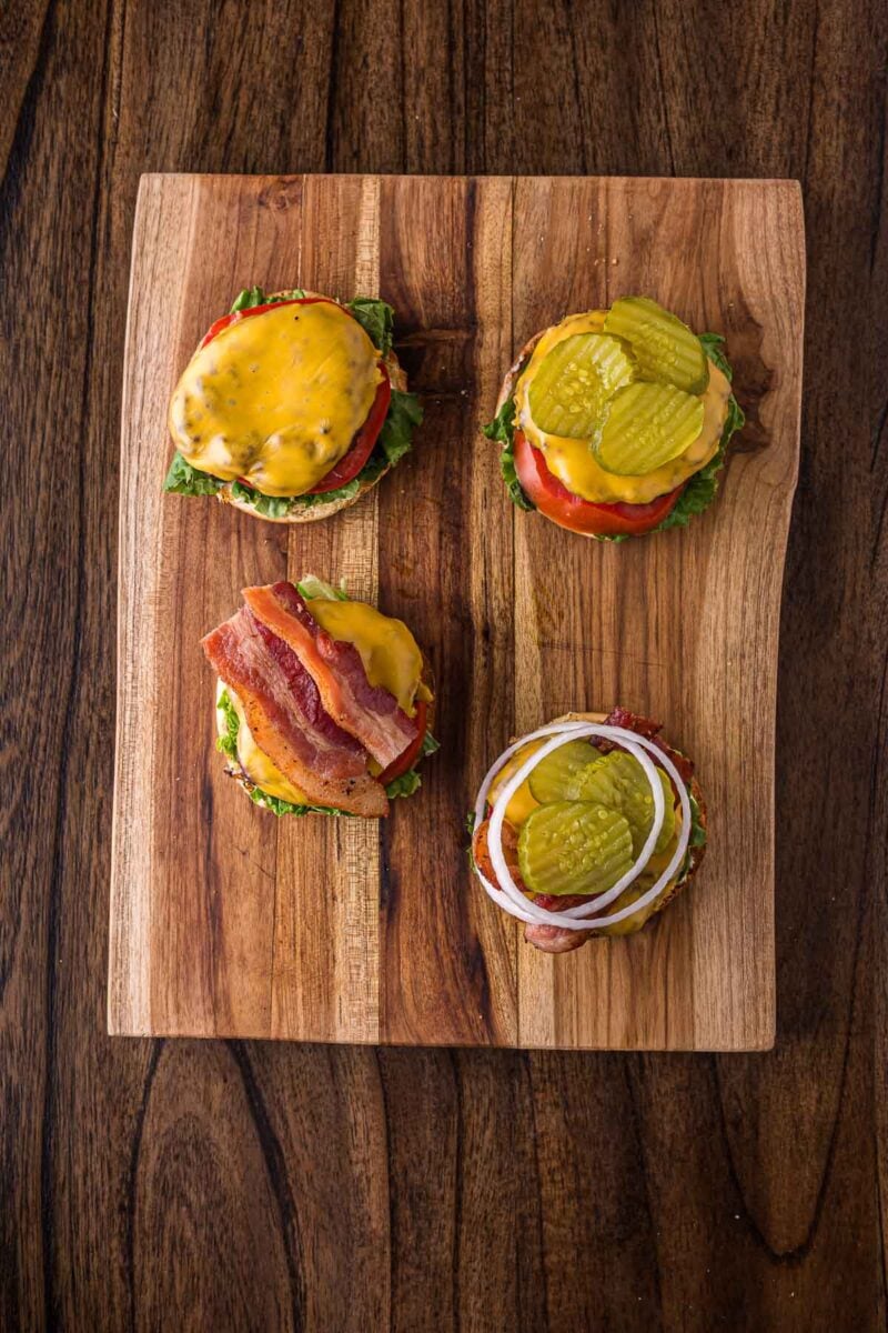 Four open-faced burgers with various toppings—cheese, bacon, pickles, tomato, lettuce, and onion—arranged on a wooden cutting board.