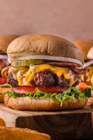 A cheeseburger with lettuce, tomato, bacon, pickles, onion, and melted cheese on a bun, placed on a wooden surface.