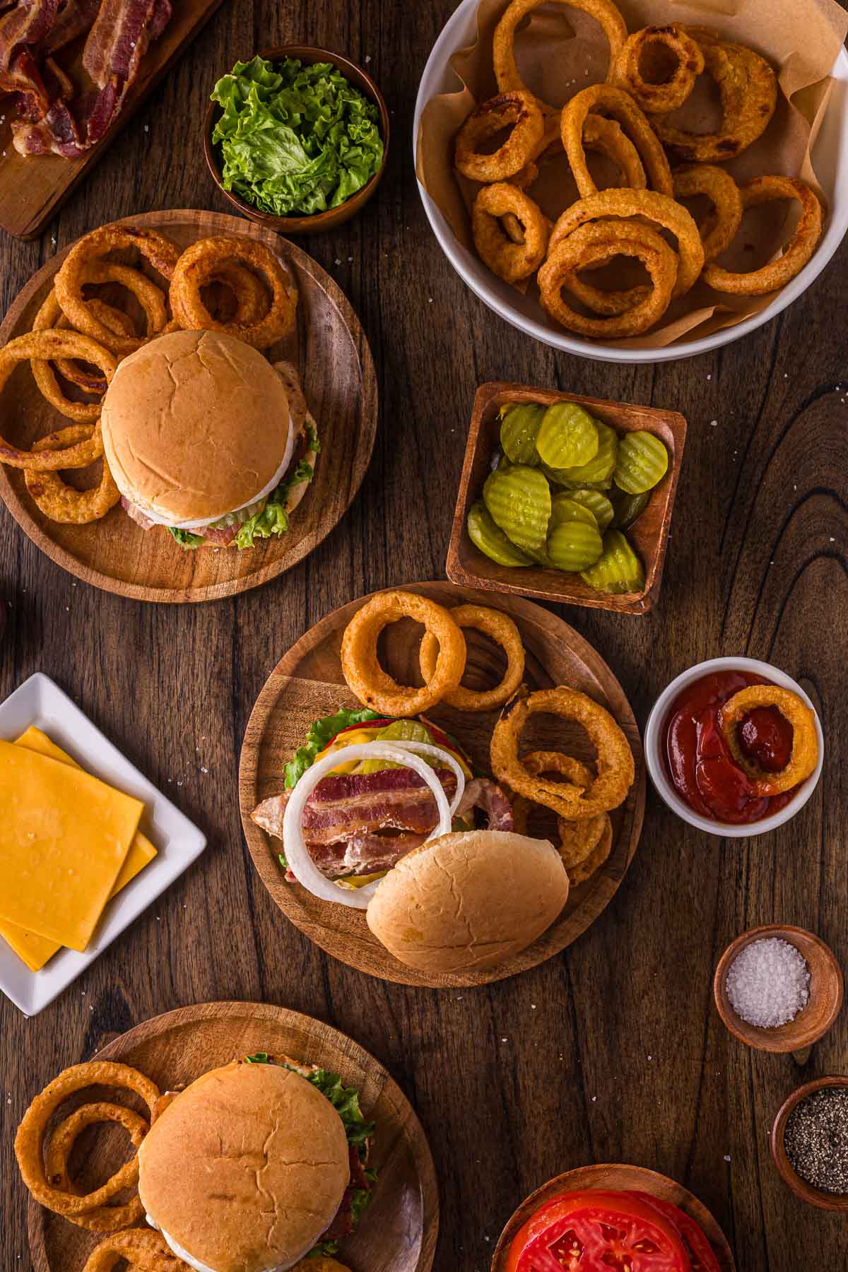 Wooden table with plates of burgers, onion rings, bacon, sliced cheese, lettuce, sliced pickles, tomato, ketchup, and small bowls of salt and pepper arranged for a meal.