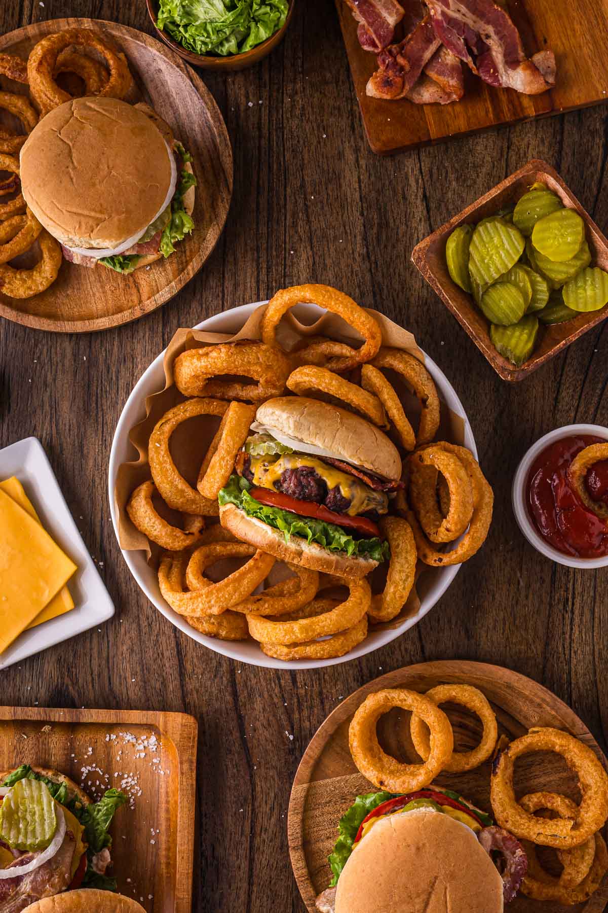 A table set with classic bacon cheeseburgers, curly fries, sliced pickles, bacon, lettuce, cheese, and a small cup of ketchup on wooden dishes.