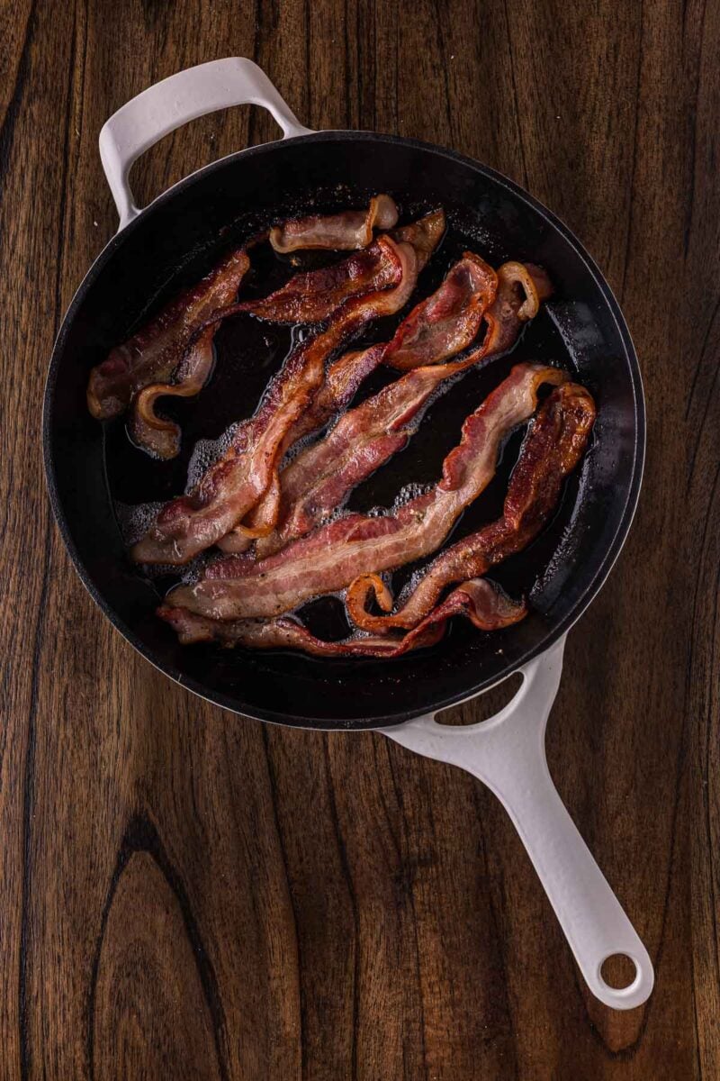 Cooked bacon strips in a white skillet on a wooden surface, viewed from above.