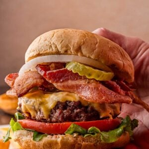 A hand holds a cheeseburger with lettuce, tomato, bacon, pickles, onion, and melted cheese, with onion rings in the background.