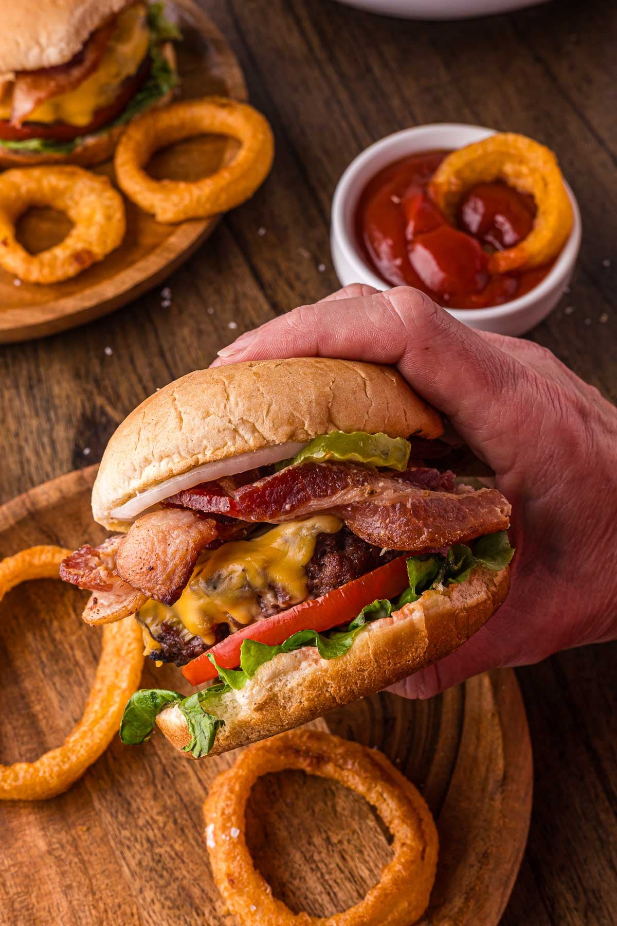 A hand holding a bacon cheeseburger with lettuce and tomato, surrounded by onion rings on a wooden plate, with a bowl of ketchup and onion rings in the background.