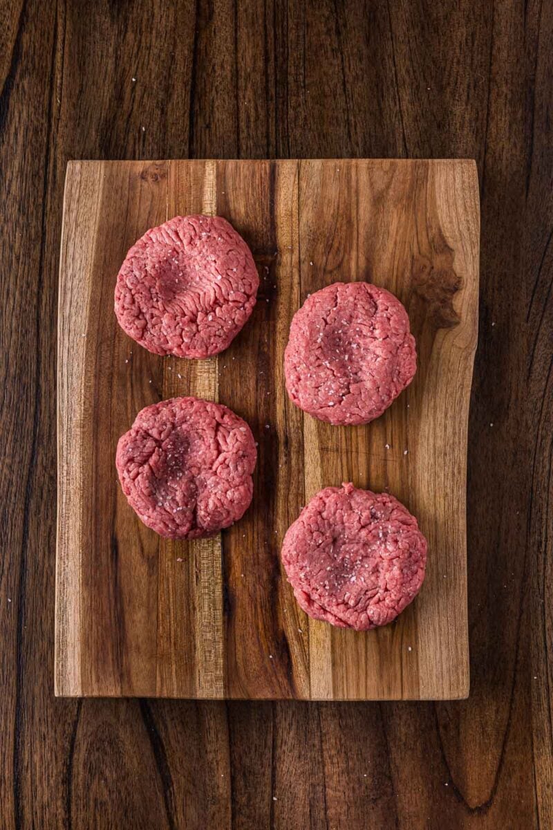 Four raw ground beef patties with a dimple in the enter of each placed on a wooden cutting board, set on a dark wood surface.