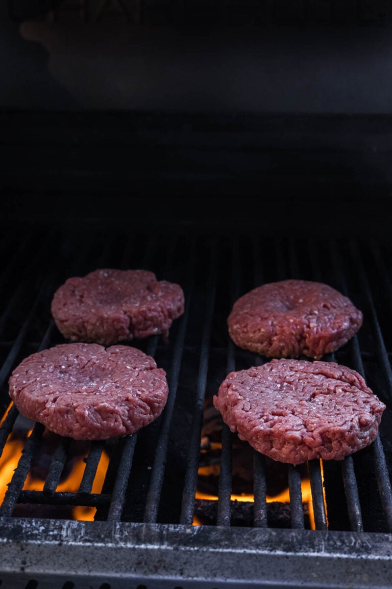 Four raw beef burger patties are cooking on a grill over open flames.