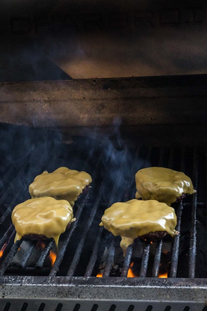 Four beef patties topped with melted cheese are cooking on a grill with visible grill marks and rising smoke.