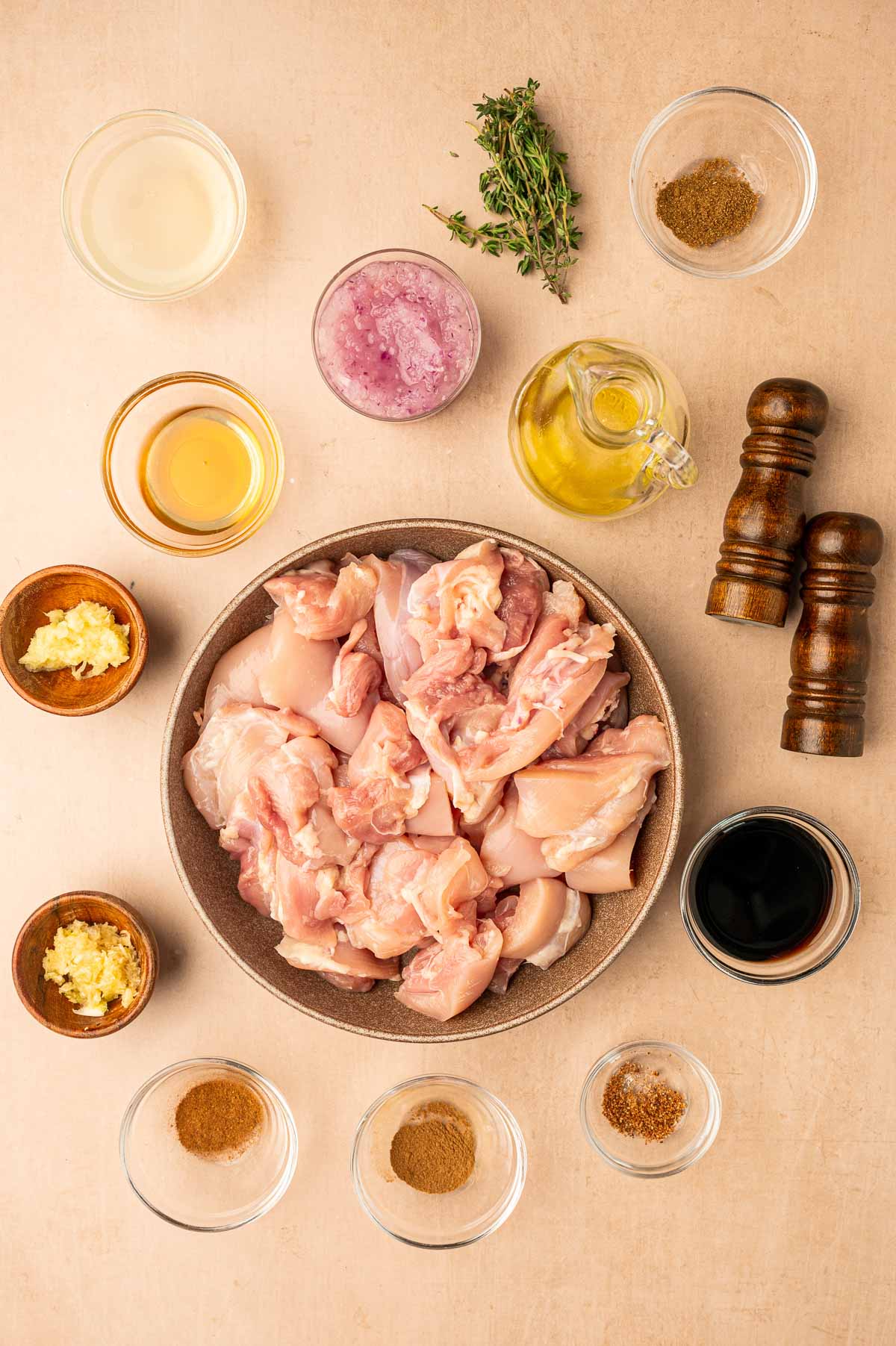 A bowl of raw chicken pieces surrounded by various herbs, spices, sauces, and condiments in small bowls, arranged on a beige surface.