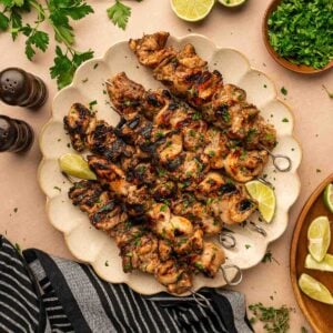 A plate of grilled jerk chicken skewers garnished with herbs, served with lime wedges. Fresh parsley, lime halves, and seasonings are arranged around the plate.