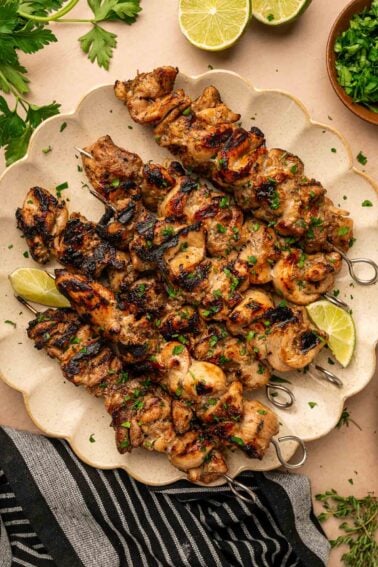 Grilled jerk chicken skewers garnished with chopped herbs are arranged on a plate, with lime wedges and fresh parsley nearby.