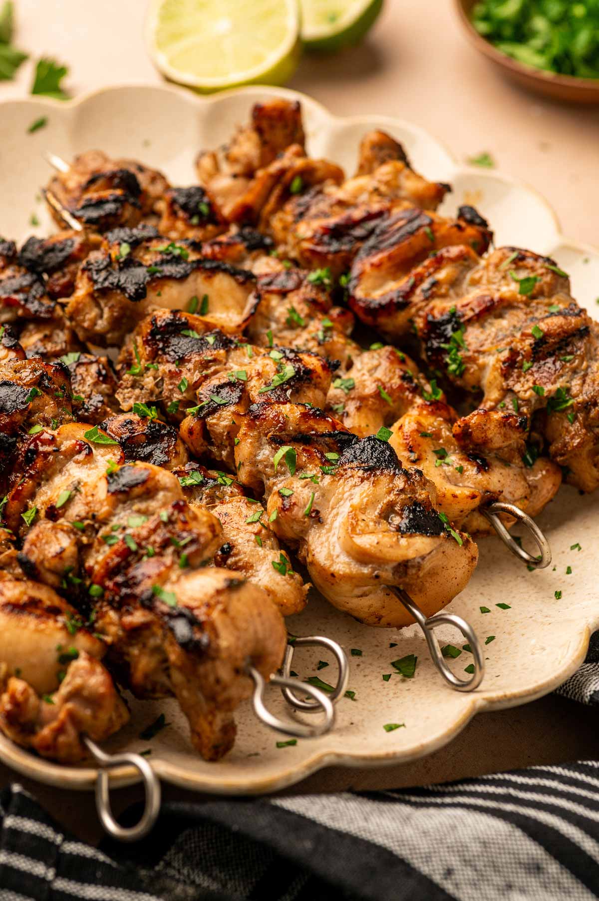 Grilled jerk chicken skewers on metal sticks arranged on a plate, garnished with chopped herbs. Lime slices and chopped greens are visible in the background.