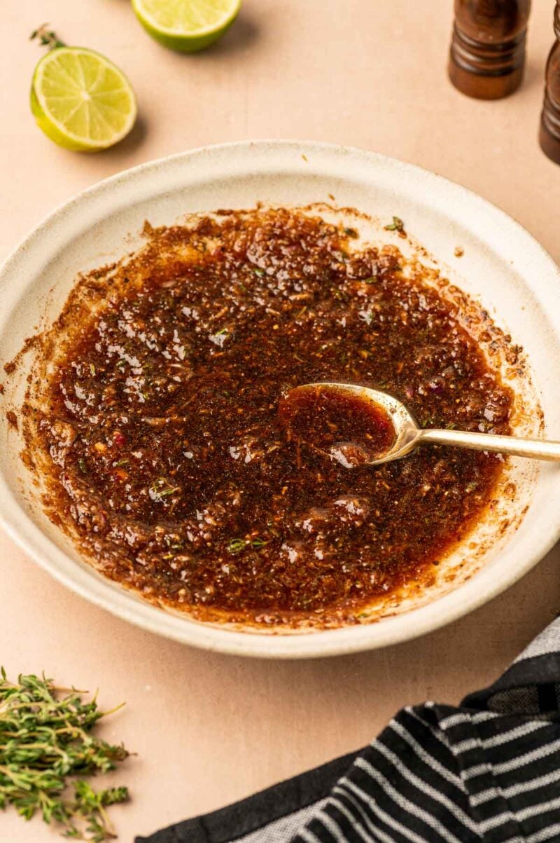 A bowl of dark, seasoned jerk marinade with a spoon, surrounded by fresh herbs, a striped cloth, and halved limes on a light surface.