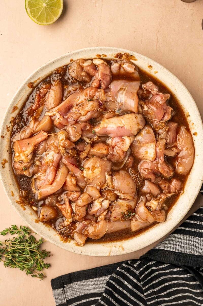 Raw chicken pieces marinating in a brown jerk marinade in a bowl, with fresh thyme, a striped cloth, and a lime half nearby on a beige surface.