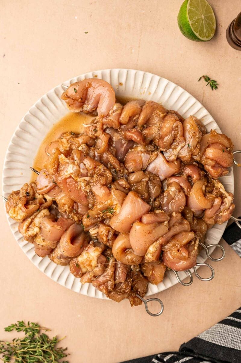 Raw marinated chicken pieces threaded onto metal skewers, arranged on a white plate, with a halved lime and herbs nearby.