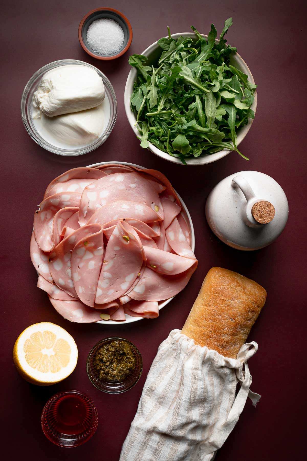 A loaf of ciabatta, slices of mortadella, arugula, mozzarella, a lemon half, a small bowl of pesto, olive oil, burrata, and a dish of salt arranged on a maroon surface.