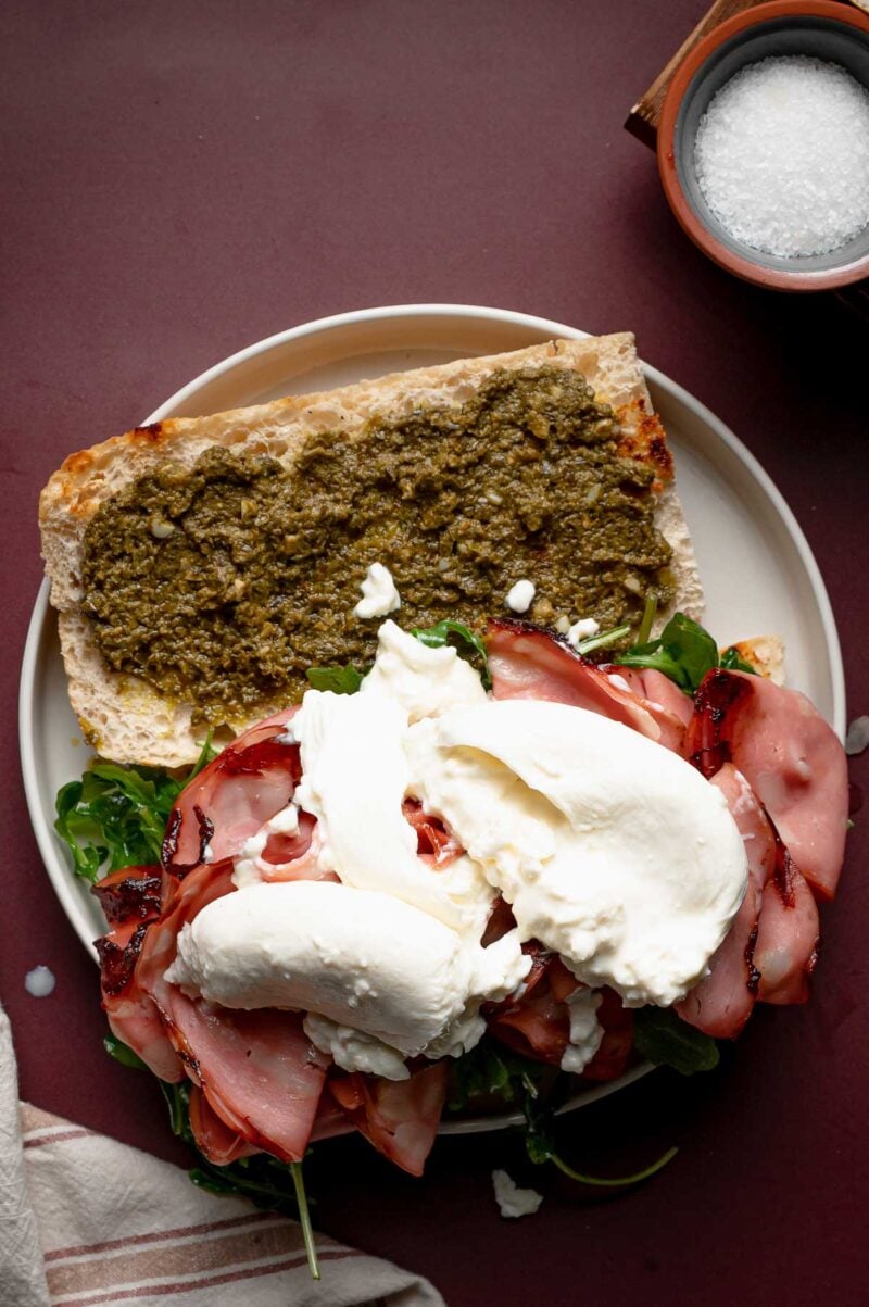 Open sandwich on a white plate with pesto spread, arugula, slices of grilled mortadella and fresh burrata cheese. A small bowl of salt and a striped cloth are nearby.