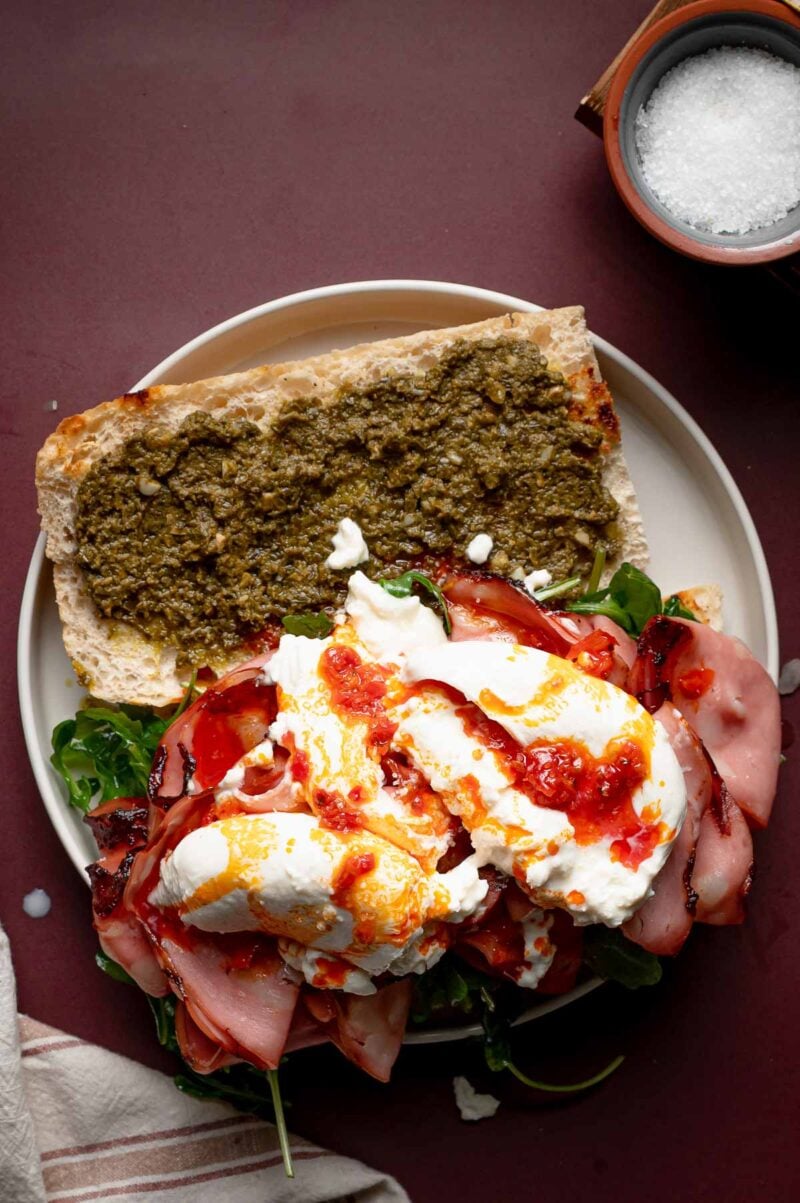 Open sandwich with pesto spread on one half of ciabatta bread, arugula, grilled mortadella, burrata cheese, and chili oil on the other half, served on a white plate.