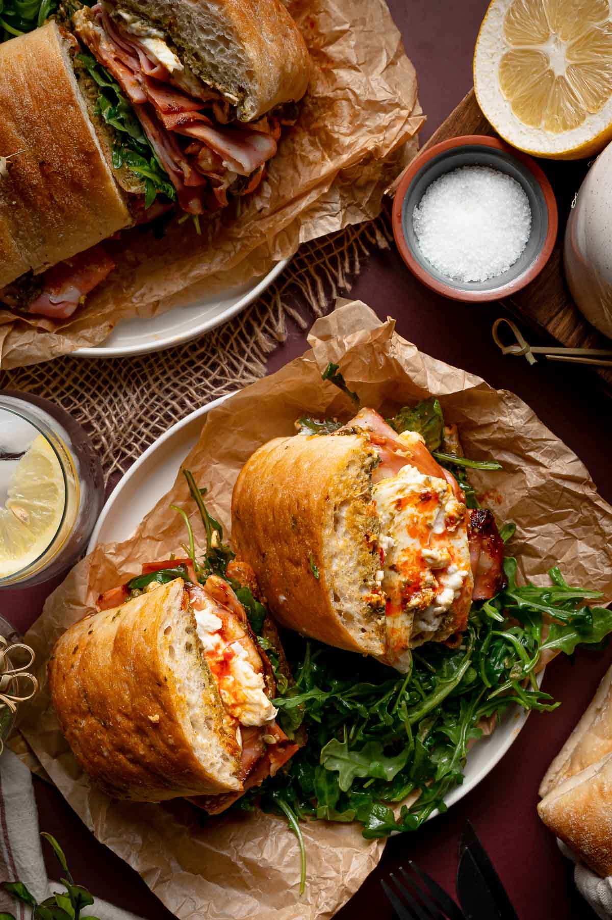 A grilled mortadella sandwich with arugula and cheese sits on brown parchment paper, with a cut lemon, salt bowl, and a glass of water with lemon nearby.