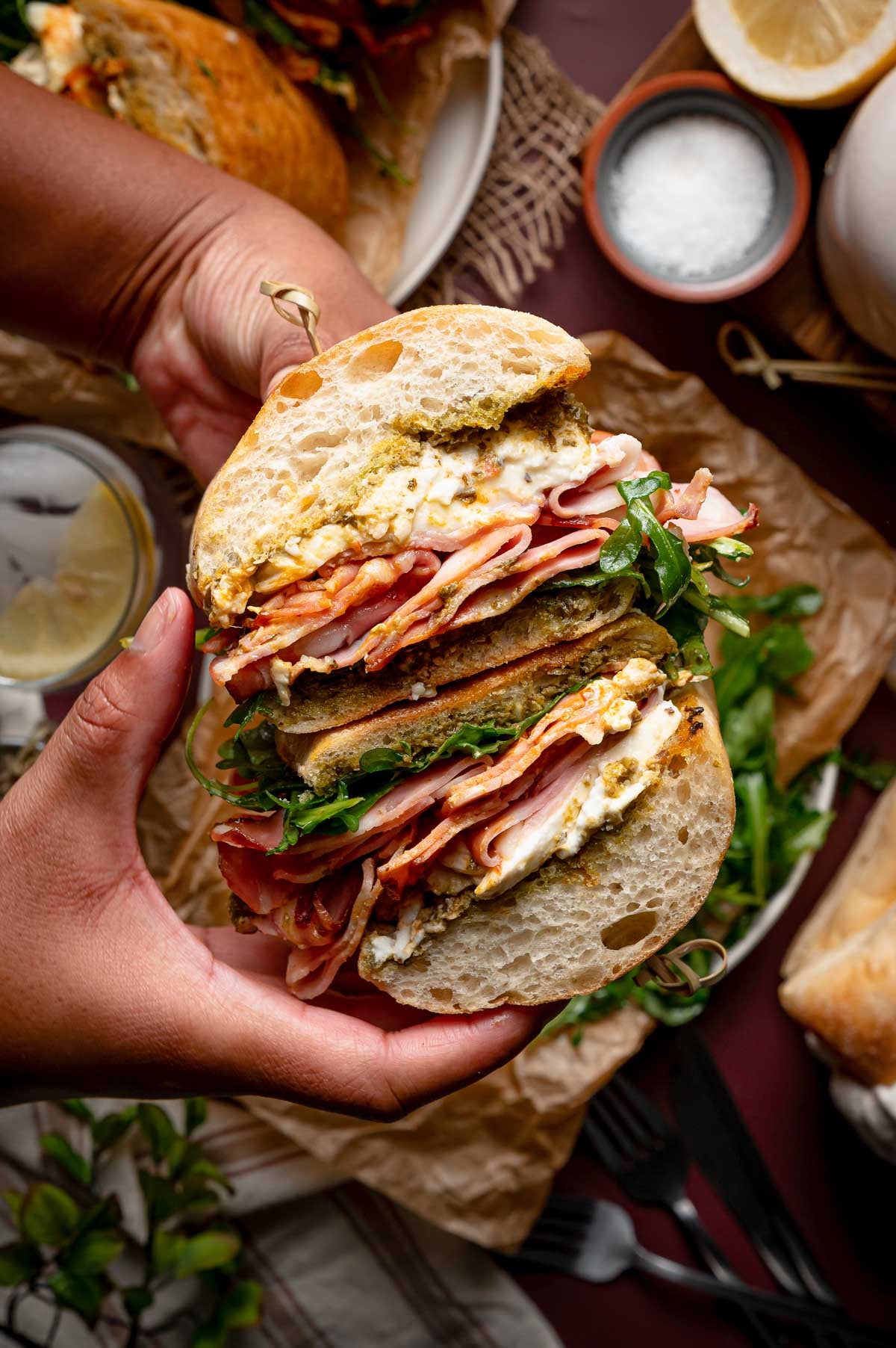 A person holds a sandwich filled with grilled mortadella, burrata, arugula, and pesto on ciabatta bread, with a lemon wedge, salt, and another sandwich visible in the background.