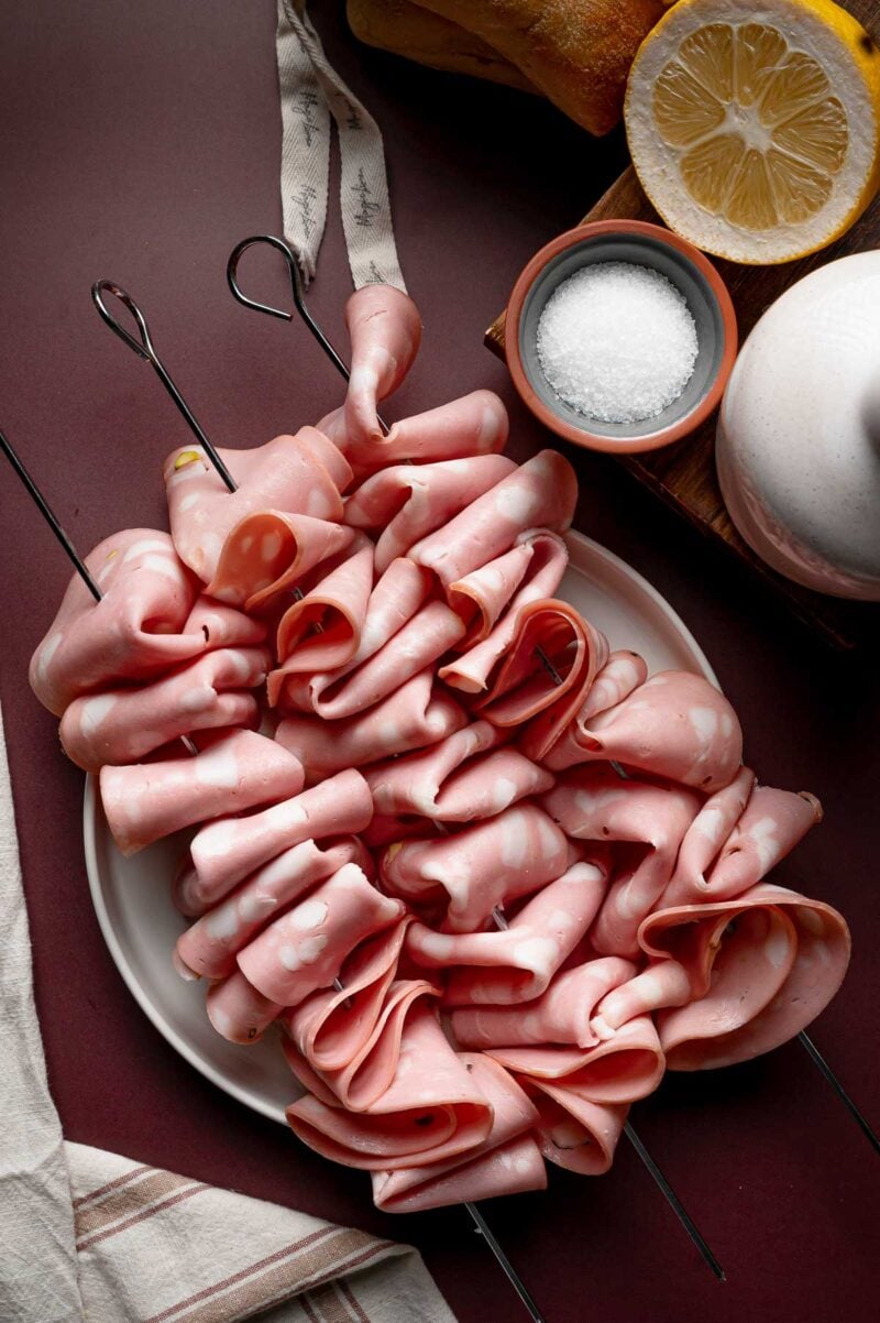 Skewers of folded mortadella are arranged on a white plate, with a bowl of salt, a halved lemon, and a loaf of bread nearby on a burgundy surface.