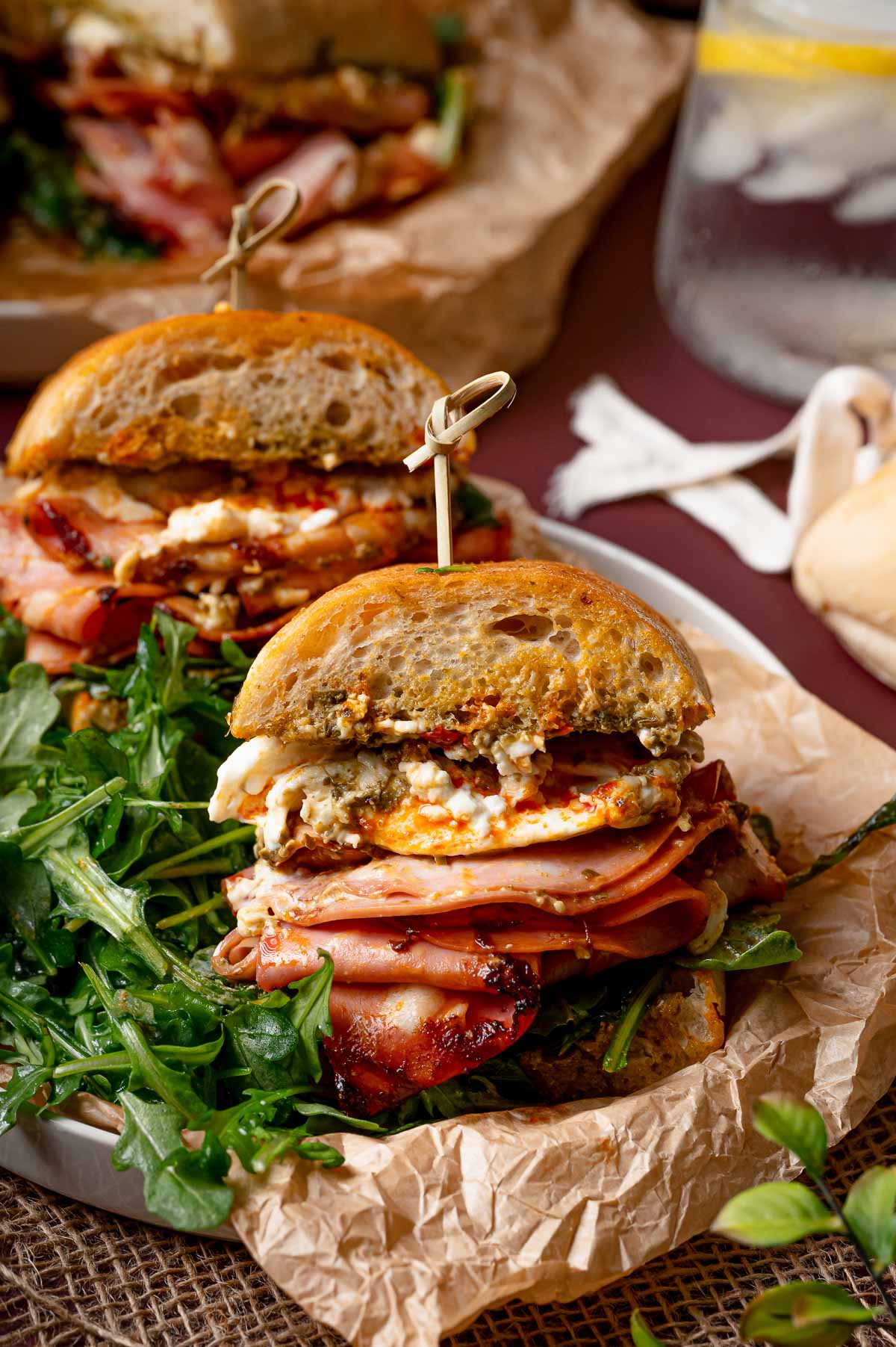 A sandwich filled with mortadella, burrata, and greens is cut in half and secured with skewers, served on parchment paper with a glass of water in the background.