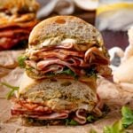 A stacked sandwich with layers of sliced grilled mortadella, burrata, greens, and pesto on crusty bread, cut in half and placed on parchment paper.