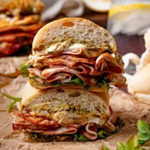 A stacked sandwich with layers of sliced grilled mortadella, burrata, greens, and pesto on crusty bread, cut in half and placed on parchment paper.