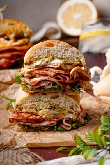 A stacked sandwich with layers of sliced grilled mortadella, burrata, greens, and pesto on crusty bread, cut in half and placed on parchment paper.