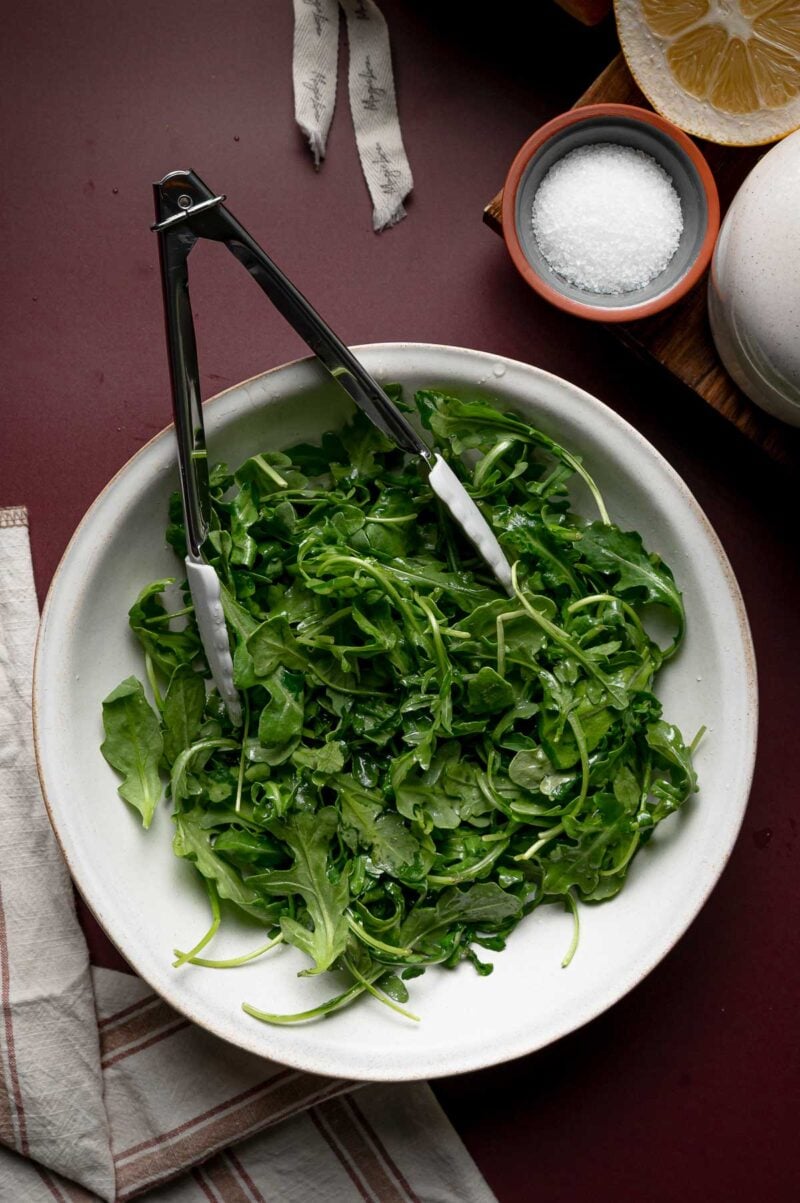 A white bowl filled with fresh arugula leaves and metal tongs, placed on a dark surface with a bowl of salt, a lemon half, and a napkin nearby.