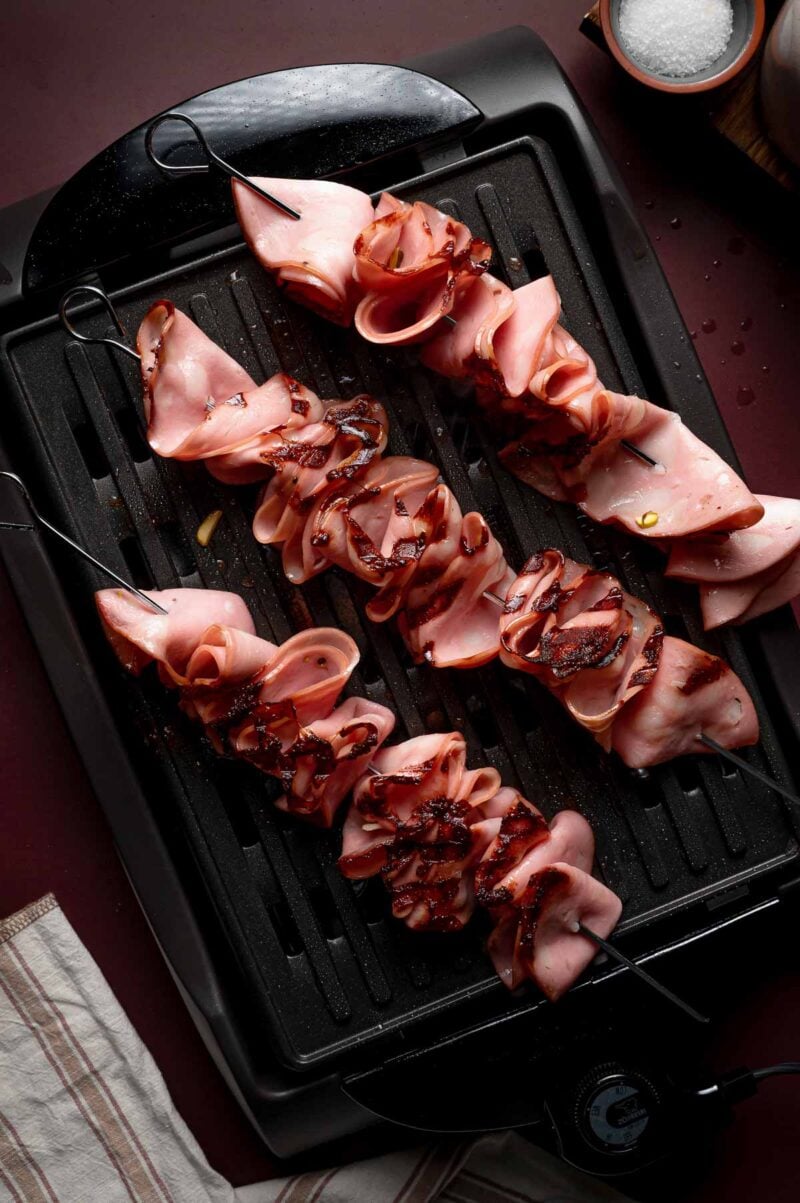 Grilling skewers of folded sliced mortadella are grilling on an electric grill, with brown edges and crispy grill marks visible.
