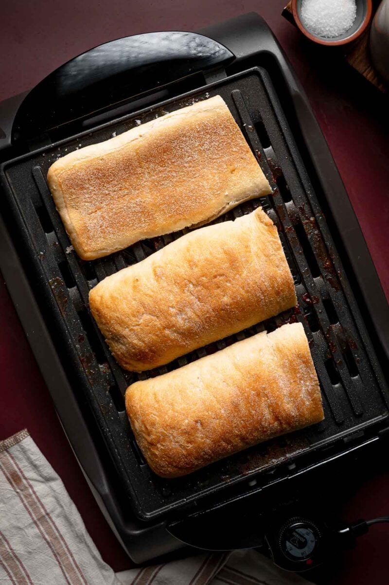 Three rectangular ciabatta loaves are toasting on an electric griddle, viewed from above.