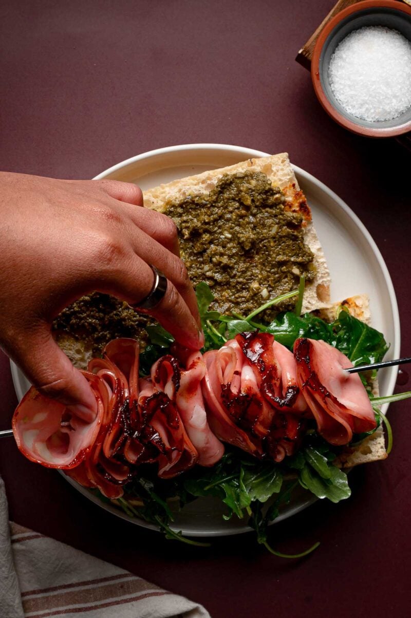 A hand assembling a sandwich with sliced grilled mortadella being pulled off a skewer, arugula, and pesto on ciabatta bread, next to a small bowl of salt.