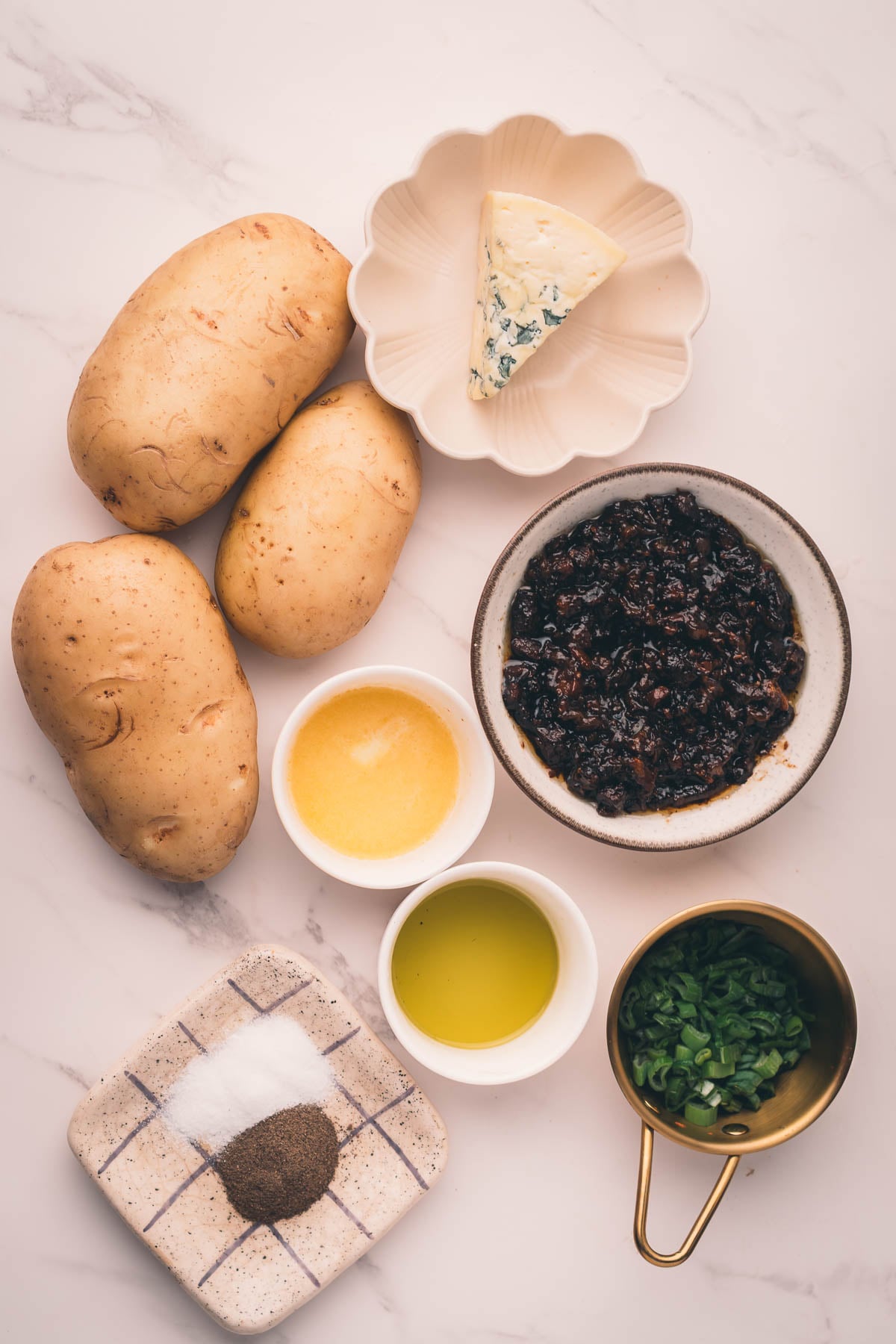 Various ingredients arranged on a white surface, including three potatoes, a wedge of cheese, bacon jam, chopped green onions, melted butter, olive oil, salt, and pepper.