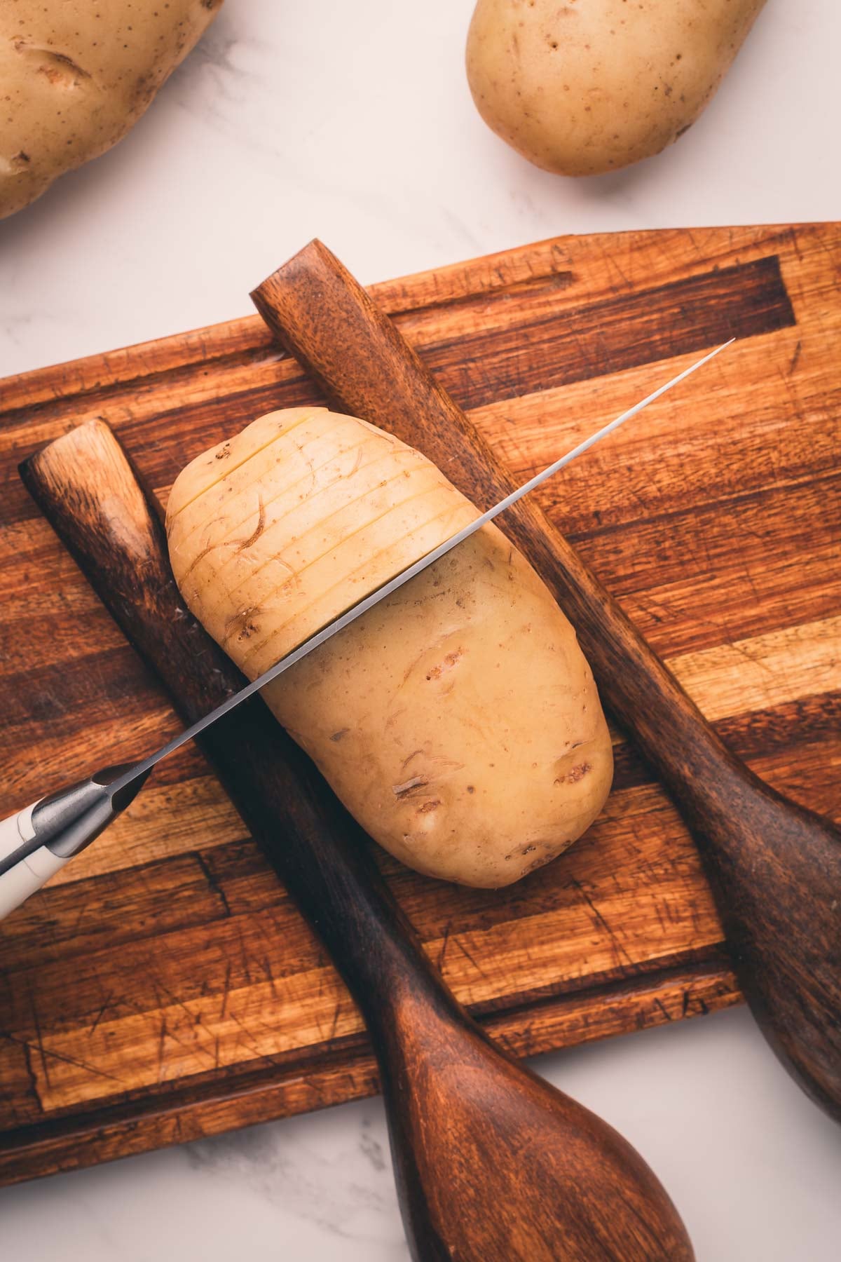 A potato is being sliced with a knife on a wooden cutting board, supported by two wooden spoons on either side so to not cut entirely through.