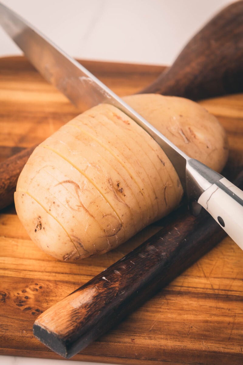 A potato with shallow, evenly spaced cuts is being sliced with a large knife on a wooden cutting board.