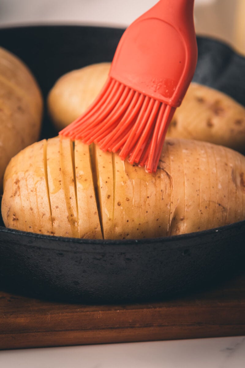 A red silicone brush applies a butter and oil mixture to sliced potatoes in a black cast iron pan.
