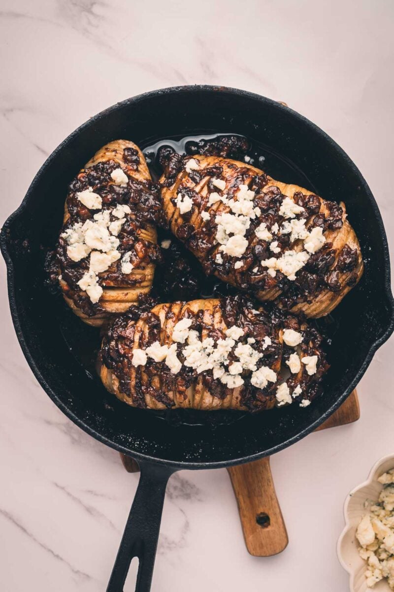 Three Hasselback potatoes with bacon jam and crumbled cheese on top, cooked in a black cast iron skillet placed on a wooden board.