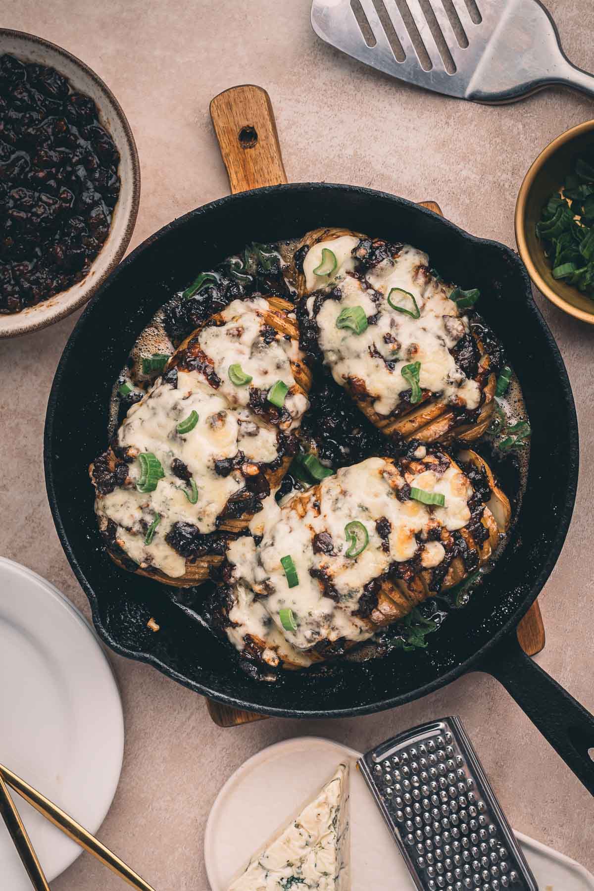 A cast iron skillet with three baked hasselback potatoes topped with melted cheese and green onions, surrounded by plates, a cheese grater, and small bowls of garnish and sauce.