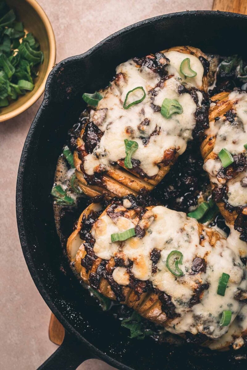 Three Hasselback potatoes topped with bacon jam, melted cheese and chopped green onions in a cast iron skillet, with a bowl of sliced green onions nearby.