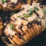 Close-up of a Hasselback potato topped with melted cheese, bacon jam, and sliced green onions in a cast iron skillet.