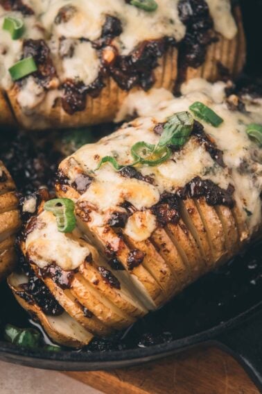 Close-up of a Hasselback potato topped with melted cheese, bacon jam, and sliced green onions in a cast iron skillet.