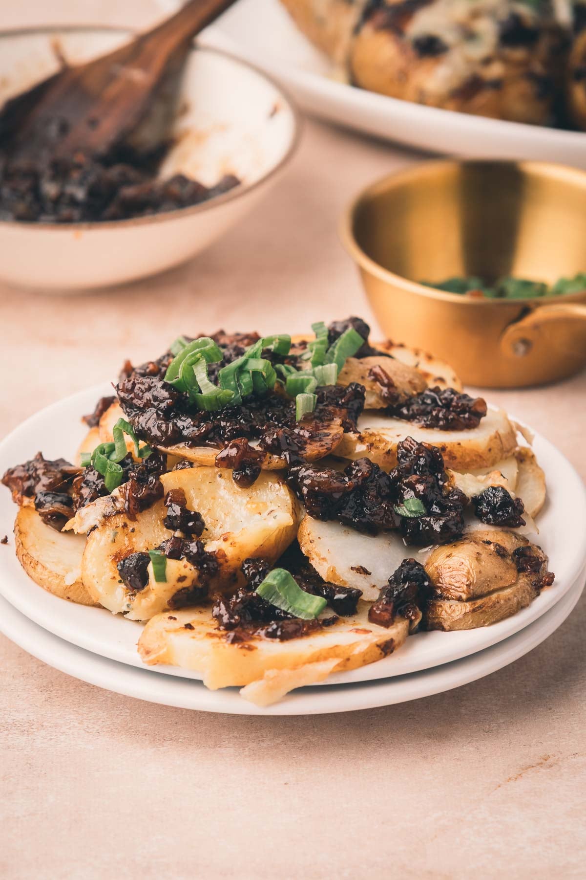 A plate of sliced baked potatoes topped with bacon jam and chopped green onions, with bowls of sauce and green onions in the background.
