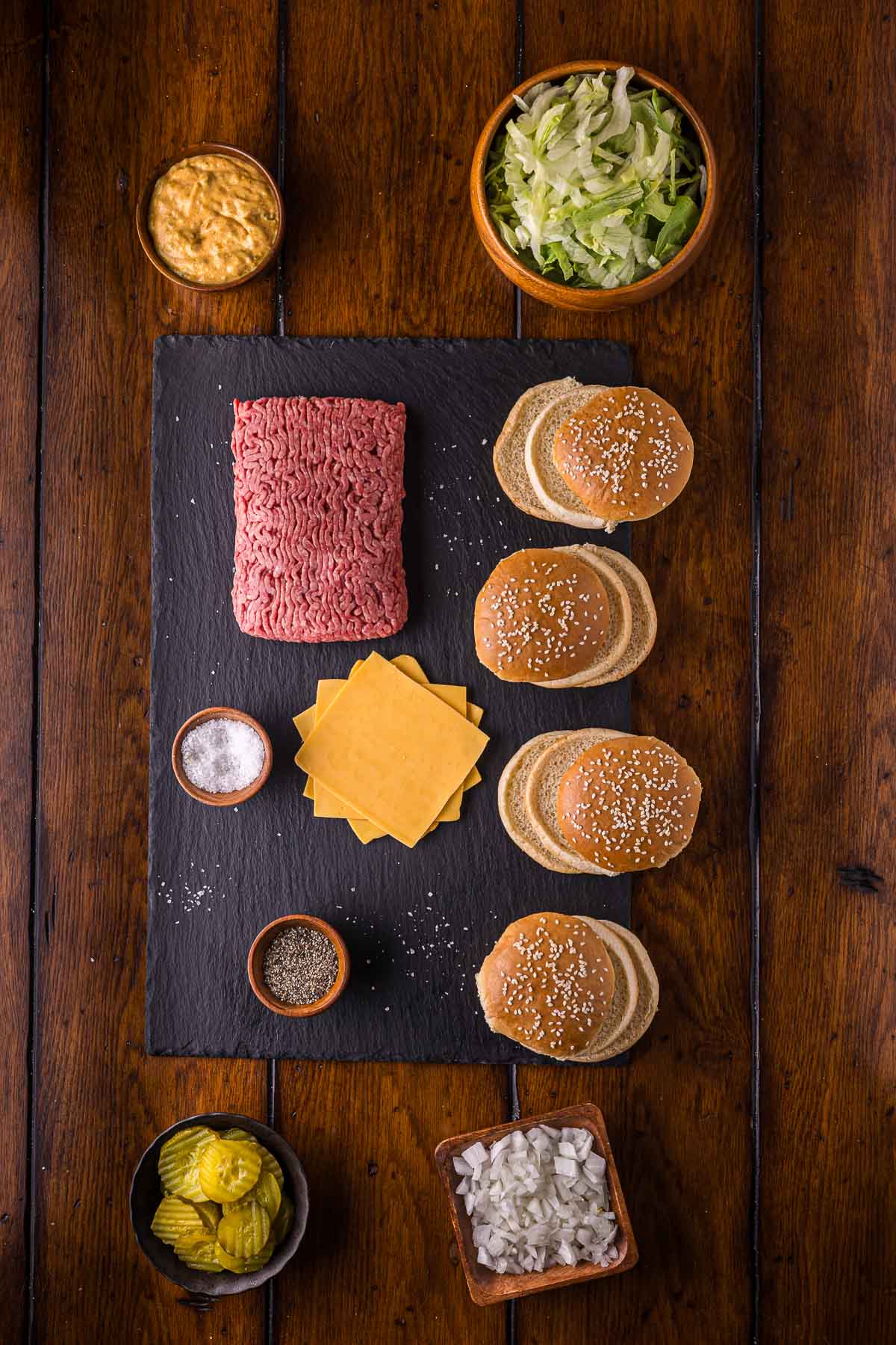 A top-down view of burger ingredients: raw ground beef, sliced cheddar cheese, sesame seed buns, shredded lettuce, pickles, diced onions, sauce, salt, and pepper on a wooden table.