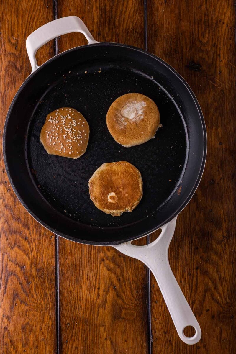 Three burger buns, one with sesame seeds, are toasting in a white-handled black skillet on a wooden surface.
