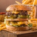 A double cheeseburger with lettuce and sauce on a wooden board, served with a side of French fries and a small container of ketchup.